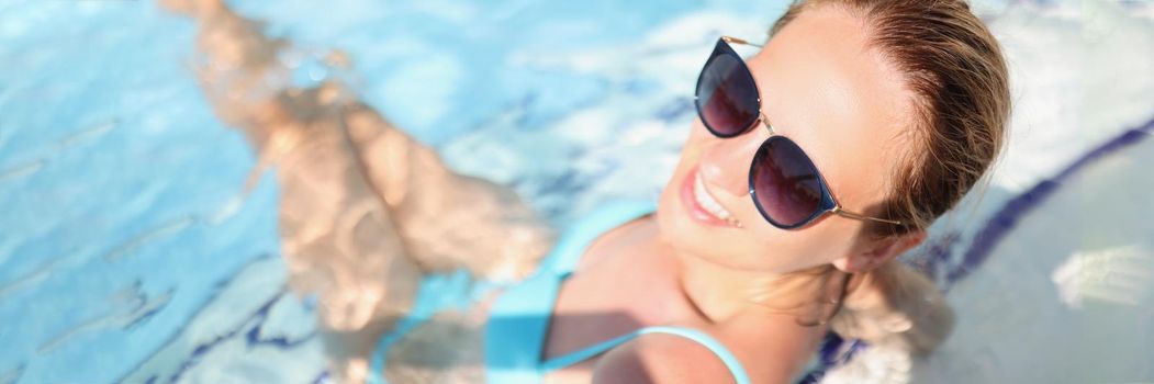 Portrait of carefree young woman relaxing in luxury swimming pool, smiling female wear glasses, blue swimsuit, clean water. Holiday, summer, resort, chilling, vacation concept