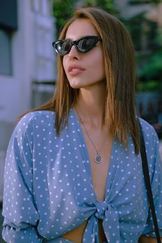 Charming blonde female in a long blue dress with polka-dots, watch, sunglasses, with a pendant around her neck and a small black handbag on her shoulder is looking away while walking alone in the city. The concept of fashion and style. Close-up shot.