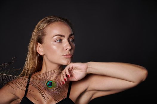 Close-up gorgeous stunning middle aged blonde Caucasian woman with healthy fresh clean skin and wet blond hair holding a peacock feather, isolated over black background with copy ad space