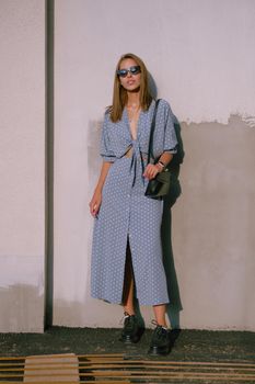 Charming blonde lady in a long blue dress with polka-dots, black boots, watch, sunglasses, with a pendant around her neck and a small handbag on her shoulder is posing near a white wall while walking alone in the city. The concept of fashion and style. Full length shot.