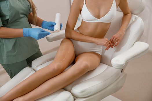 Cosmetologist applying ointment on client legs before photo epilation procedure. Hair removal