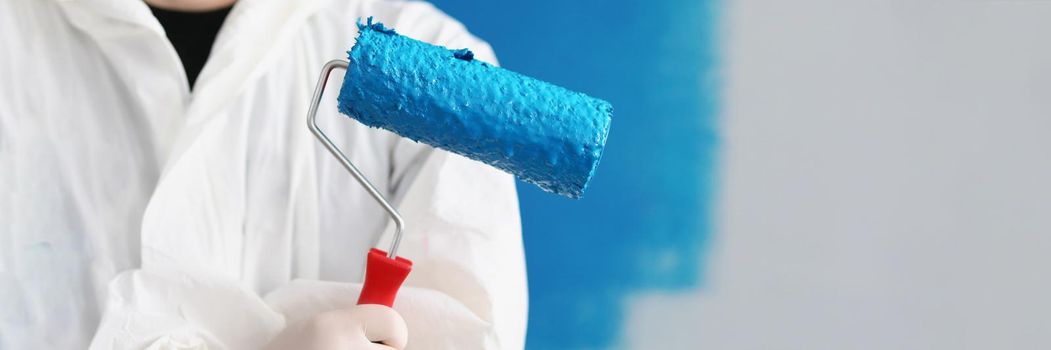 Close-up of painter man hand holding roller tool for painting walls, blue colour on equipment, worker in uniform. Copy space in right. Renovation, interior design, redecoration concept