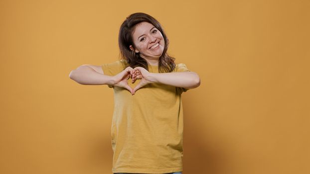 Happy woman gesturing making heart sign with hands to show loving feelings for her partner over yellow background. Smiling person feeling in love talking romantic for valentine day.