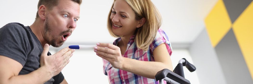 Low angle of woman in wheelchair show to husband positive pregnancy test. Surprised reaction of man, bright emotion. Disabled people, family growing, happiness, healthcare concept