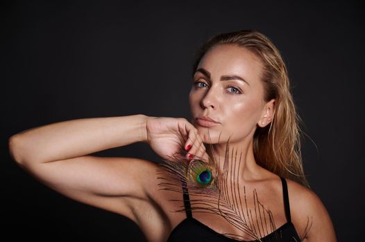 Attractive middle aged blonde woman with healthy fresh clean skin holding a peacock feather, isolated over black background with copy ad space