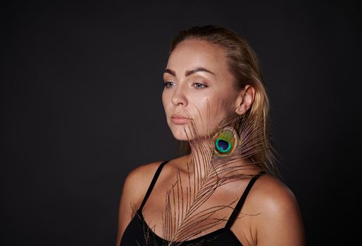 Attractive middle aged blonde woman with healthy fresh clean tanned radiant skin holding a peacock feather, isolated over black background with copy ad space