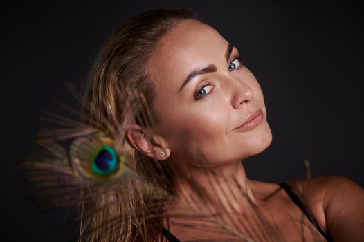 Headshot gorgeous smiling stunning middle aged blonde Caucasian woman with healthy fresh clean skin and wet blond hair holding a peacock feather, isolated over black background with copy ad space