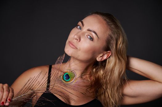 Attractive middle aged blonde woman with healthy fresh clean skin holding a peacock feather, isolated over black background with copy ad space