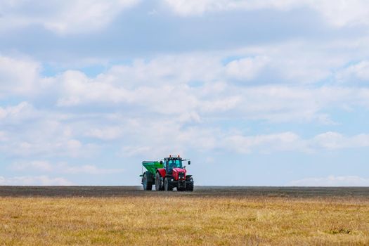 A red tractor in a green trailer is carrying fertilizer to the field. Agricultural work is performed by a farmer.