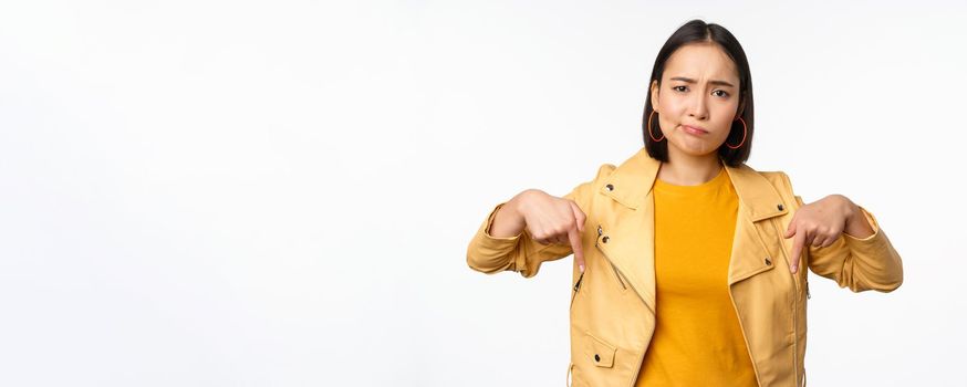 Portrait of disappointed asian woman pointing fingers down, grimacing and showing smth bad, standing over white background. Copy space