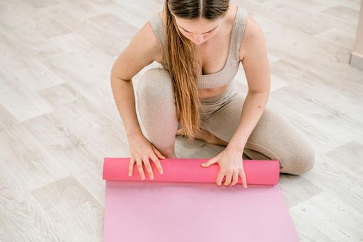 A young woman rolls a pink fitness or yoga mat before or after exercising, exercising at home in the living room or in a yoga studio. Healthy habits, keep fit, weight loss concept. Closeup photo.