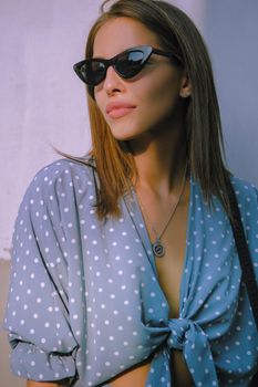 Pretty blonde female in a long blue dress with polka-dots, watch, sunglasses, with a pendant around her neck and a small black handbag on her shoulder is walking alone in the city. The concept of fashion and style. Close-up shot.