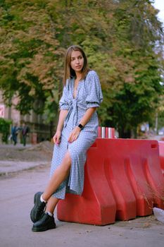 Attractive blonde girl in a long blue dress with polka-dots, black boots, watch, with a pendant around her neck and a small handbag on her shoulder is posing leaning a red guardrail while walking alone in the city. The concept of fashion and style. Full length shot.