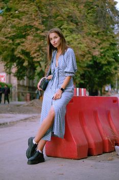 Attractive blonde lady in a long blue dress with polka-dots, black boots, watch, with a pendant around her neck and a small handbag on her shoulder is looking at the camera, posing leaning a red guardrail while walking alone in the city. The concept of fashion and style. Full length shot.