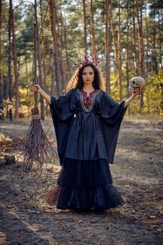 Pretty, wicked, long-haired sibyl in a black, long embroidered dress. There is large red crown in her brown, curly hair. She is holding her broom and a skull while posing in a pine forest. Spells, magic and witchcraft. Full length portrait.
