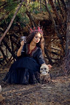 Beautiful, wicked, long-haired sibyl in a black, long embroidered dress. There is large red crown in her brown, curly hair. She is posing with her broom and a skull while sitting near a hut in a pine forest. Spells, magic and witchcraft. Full length portrait.