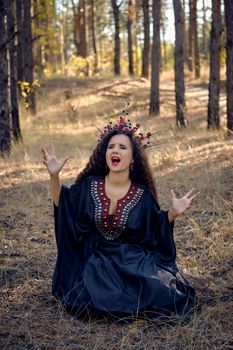 Gorgeous, wicked, long-haired sibyl in a black, long embroidered dress. There is large red crown in her brown, curly hair. She is posing sitting in a pine forest and pronouncing a spell. Magic and witchcraft. Full length portrait.