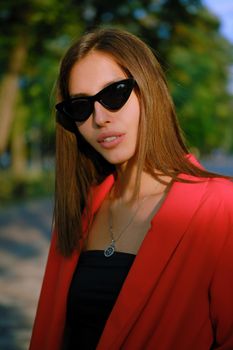 Wonderful blonde lady in a red lady-type pantsuit and black top, watch, ring, sunglasses, with a pendant around her neck is looking at the camera while walking alone in the city. The concept of fashion and style. Close-up shot.