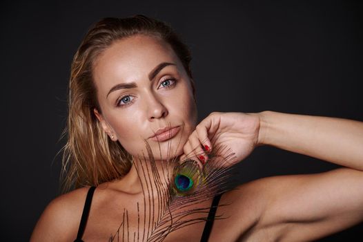 Close-up attractive middle aged blonde Caucasian woman with healthy fresh clean skin and wet blond hair holding a peacock feather, isolated over black background with copy ad space
