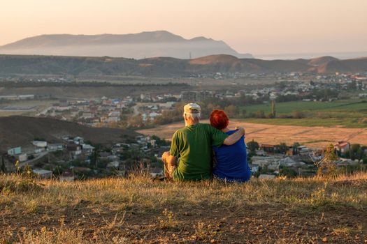 An elderly couple sits on a mountain with their backs with a beautiful view of the mountains and the sea in the distance