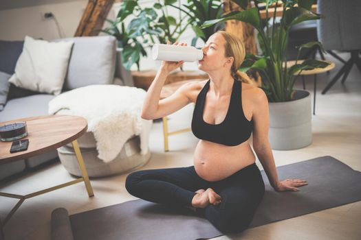 Young happy and cheerful beautiful pregnant woman taking a break, hydrating, drinking water from the botle after home well being workout progrem