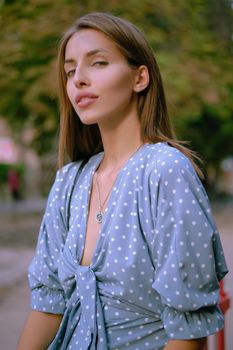 Alluring blonde female in a long blue dress with polka-dots, watch, with a pendant around her neck and a small black handbag on her shoulder is looking at the camera while walking alone in the city. The concept of fashion and style. Close-up shot.