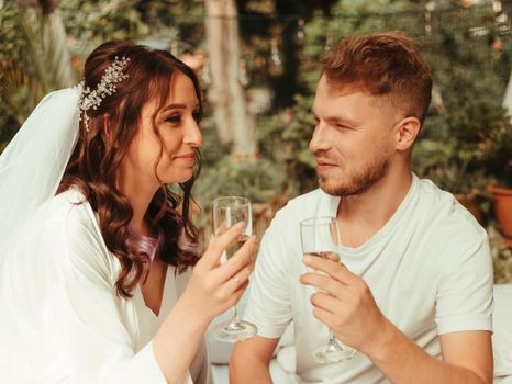 Stylish, smiling groom and cute blonde bride with curly hair in a white dress are sitting on the rug with glasses of champagne on the grass in autumn in nature. Wedding photography of the newlyweds.