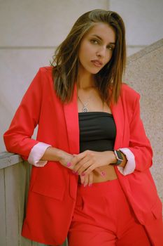 Charming blonde lady in a red lady-type pantsuit and black top, watch, ring, with a pendant around her neck is looking at the camera while walking alone in the city. The concept of fashion and style. Close-up shot.