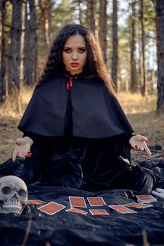Cute, wicked, long-haired sibyl in a black, long dress with cape and hood. She is sitting on a dark blanket whith fortune-telling cards and skulls on it while posing in a pine forest. Spells, magic and witchcraft. Full length portrait.
