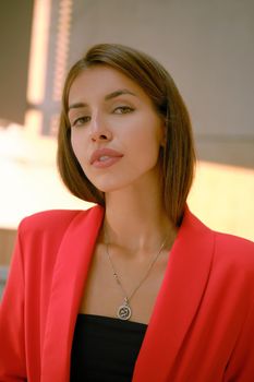 Attractive blonde lady in a red lady-type pantsuit and black top, with a pendant around her neck is looking at the camera while walking alone in the city. The concept of fashion and style. Close-up shot.