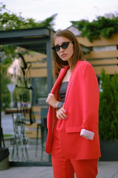 Alluring blonde woman in a red lady-type pantsuit and black top, watch, ring, sunglasses, with a pendant around her neck is walking alone in the city. The concept of fashion and style. Close-up shot.