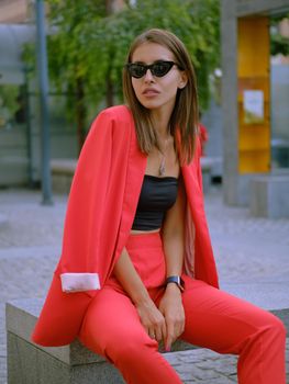 Alluring blonde lady in a red lady-type pantsuit and black top, watch, ring, sunglasses, with a pendant around her neck is posing sitting on a stone bench while walking alone in the city. The concept of fashion and style. Close-up shot.