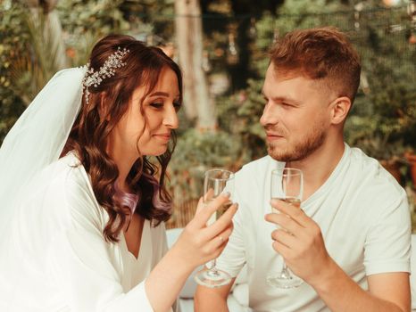 Stylish, smiling groom and cute blonde bride with curly hair in a white dress are sitting on the rug with glasses of champagne on the grass in autumn in nature. Wedding photography of the newlyweds.