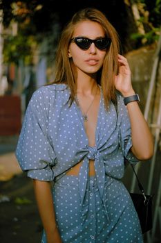 Elegant blonde lady in a long blue dress with polka-dots, watch, sunglasses, with a pendant around her neck and a small black handbag on her shoulder is walking alone in the city. The concept of fashion and style. Close-up shot.