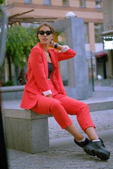 Pretty blonde woman in a red lady-type pantsuit and black top, boots, watch, ring, sunglasses, with a pendant around her neck is posing sitting sideways on a stone bench while walking alone in the city. The concept of fashion and style. Full-length shot.