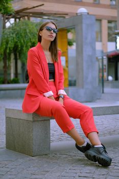 Pretty blonde female in a red lady-type pantsuit and black top, boots, watch, ring, sunglasses, with a pendant around her neck is looking up, posing sitting sideways on a stone bench while walking alone in the city. The concept of fashion and style. Full-length shot.