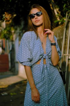 Elegant blonde maiden in a long blue dress with polka-dots, watch, sunglasses, with a pendant around her neck and a small black handbag on her shoulder is walking alone in the city. The concept of fashion and style. Close-up shot.