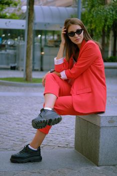 Cute blonde lady in a red lady-type pantsuit and black top, boots, watch, ring, sunglasses, with a pendant around her neck has crossed her legs and posing sitting sideways on a stone bench while walking alone in the city. The concept of fashion and style. Full-length shot.