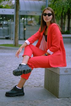 Cute blonde girl in a red lady-type pantsuit and black top, boots, watch, ring, sunglasses, with a pendant around her neck has crossed her legs and posing sitting sideways on a stone bench while walking alone in the city. The concept of fashion and style. Full-length shot.