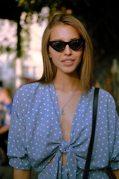 Graceful blonde female in a long blue dress with polka-dots, watch, sunglasses, with a pendant around her neck and a small black handbag on her shoulder is smiling while walking alone in the city. The concept of fashion and style. Close-up shot.