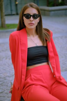 Pretty blonde girl in a red lady-type pantsuit and black top, sunglasses, with a pendant around her neck is looking at the camera, posing sitting on a stone bench while walking alone in the city. The concept of fashion and style. Close-up shot.