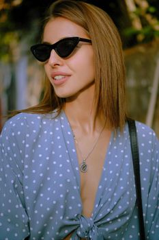 Graceful blonde girl in a long blue dress with polka-dots, watch, sunglasses, with a pendant around her neck and a small black handbag on her shoulder is smiling and looking away while walking alone in the city. The concept of fashion and style. Close-up shot.