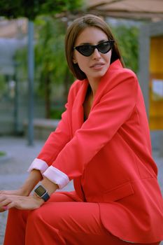 Pretty blonde maiden in a red lady-type pantsuit and black top, watch, ring, sunglasses, with a pendant around her neck is smiling, posing sitting sideways on a stone bench while walking alone in the city. The concept of fashion and style. Close-up shot.