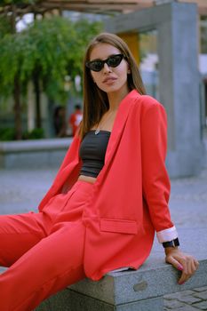 Cute blonde maiden in a red lady-type pantsuit and black top, watch, ring, sunglasses, with a pendant around her neck is posing sitting sideways on a stone bench while walking alone in the city. The concept of fashion and style. Close-up shot.
