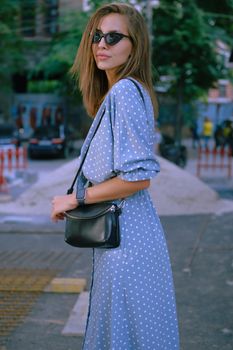 Charming blonde girl in a long blue dress with polka-dots, watch, sunglasses, with a pendant around her neck and a small black handbag on her shoulder is posing sideways while walking alone in the city. The concept of fashion and style. Close-up shot.