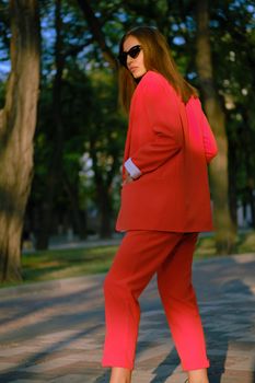 Beautiful blonde female in a red lady-type pantsuit and black top, watch, ring, sunglasses, with a pendant around her neck is posing sideways and looking away while walking alone in the city. The concept of fashion and style. Close-up shot.