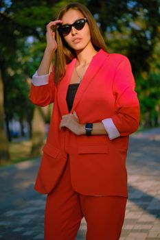 Beautiful blonde girl in a red lady-type pantsuit and black top, watch, ring, sunglasses, with a pendant around her neck is touching her hair while walking alone in the city. The concept of fashion and style. Close-up shot.