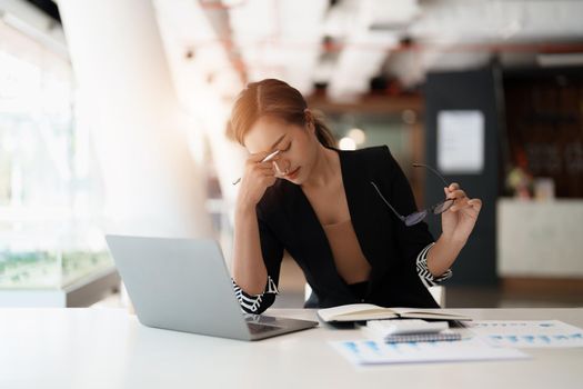 Very stressed business asian woman sitting in front of her computer looking at a large pile of paperwork, while holding a hand at her forehead.