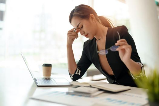 Very stressed business asian woman sitting in front of her computer looking at a large pile of paperwork, while holding a hand at her forehead.