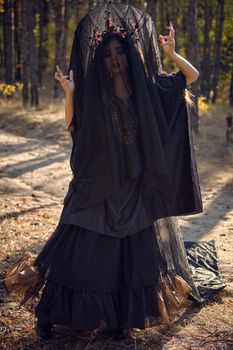 Beautiful, wicked, long-haired sibyl in a black, long embroidered dress. There is large red crown in her brown, curly hair under a black veil. She is posing in a pine forest. Spells, magic and witchcraft. Close-up.